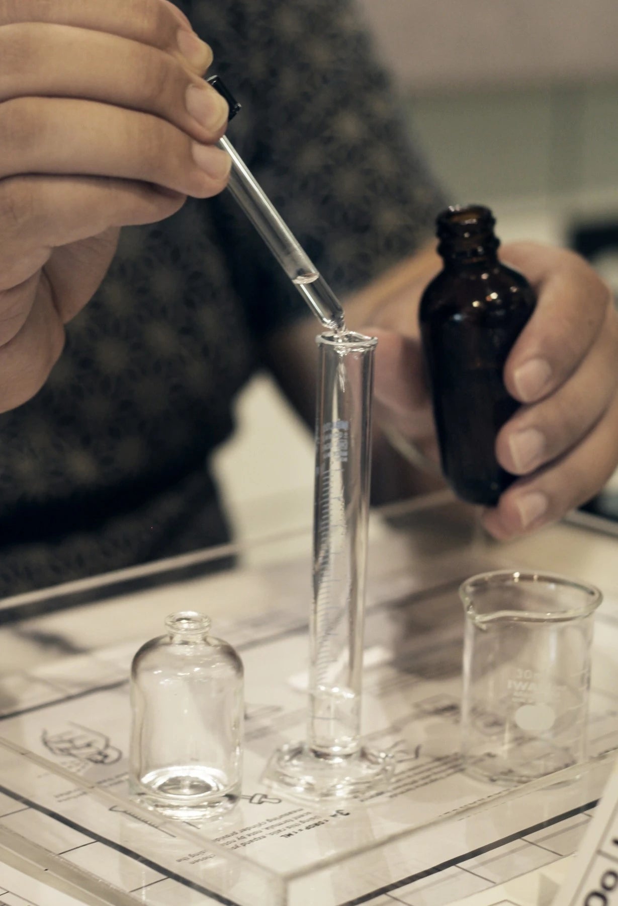 Hands blending a fragrance at the Oo La Lab workbench, using a glass pipette, measuring cylinder and fragrance notes
